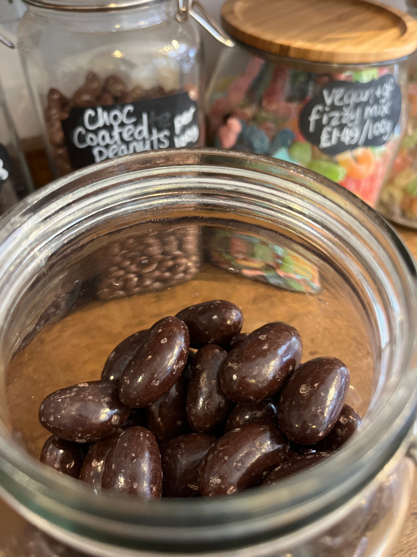 Large dark brown chocolate covered brazil nuts are just shown in an open jar in front of a row of various colourful sweet treats.