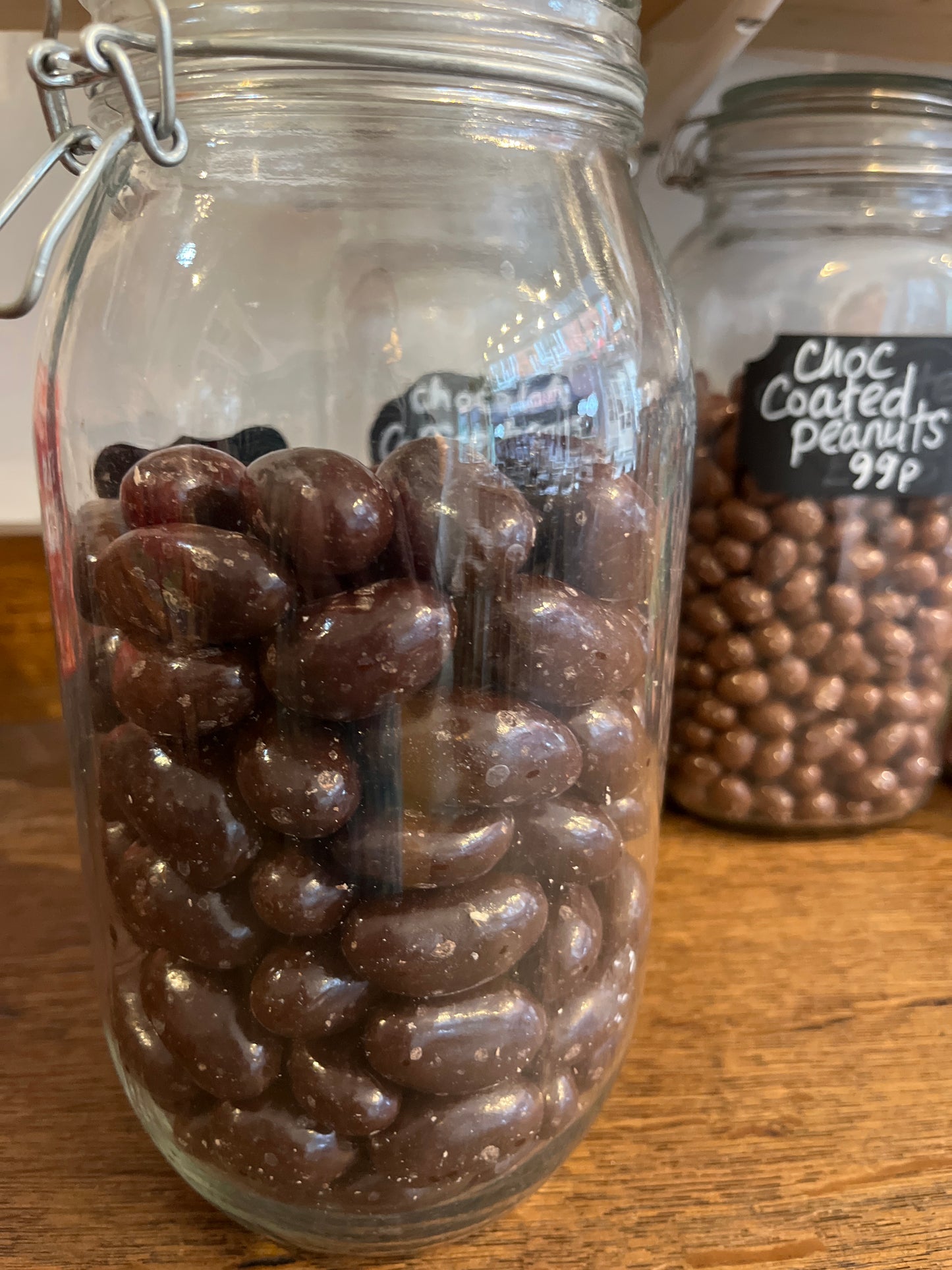 A tall jar holds large dark brown shiny ovals of chocolate coated brazil nuts. Another jar of choc covered peanuts sits on the wooden surface just behind it.